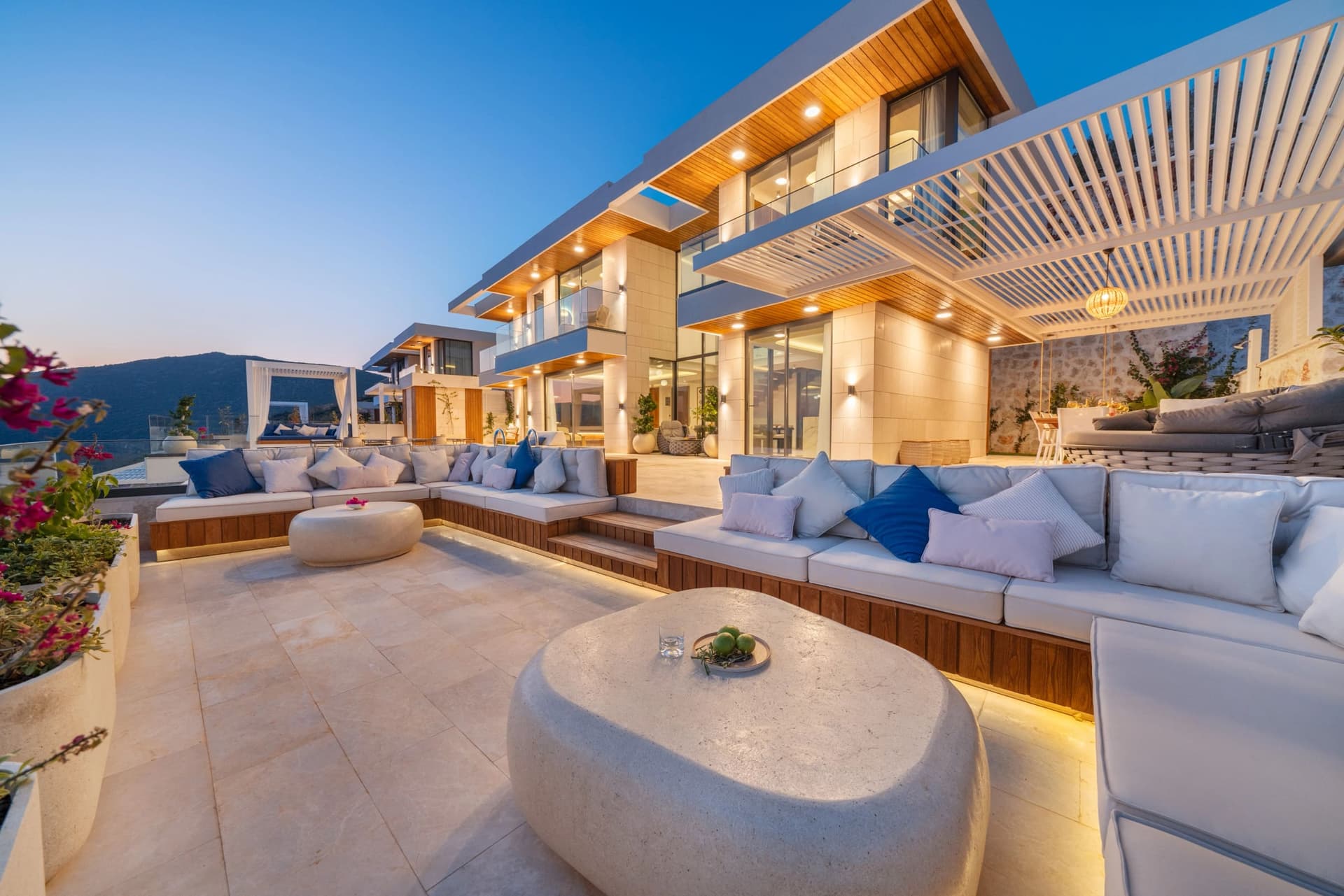 Akşam ışıklandırmalı, geniş oturma grupları ve taş masalarla döşenmiş modern villa terası.