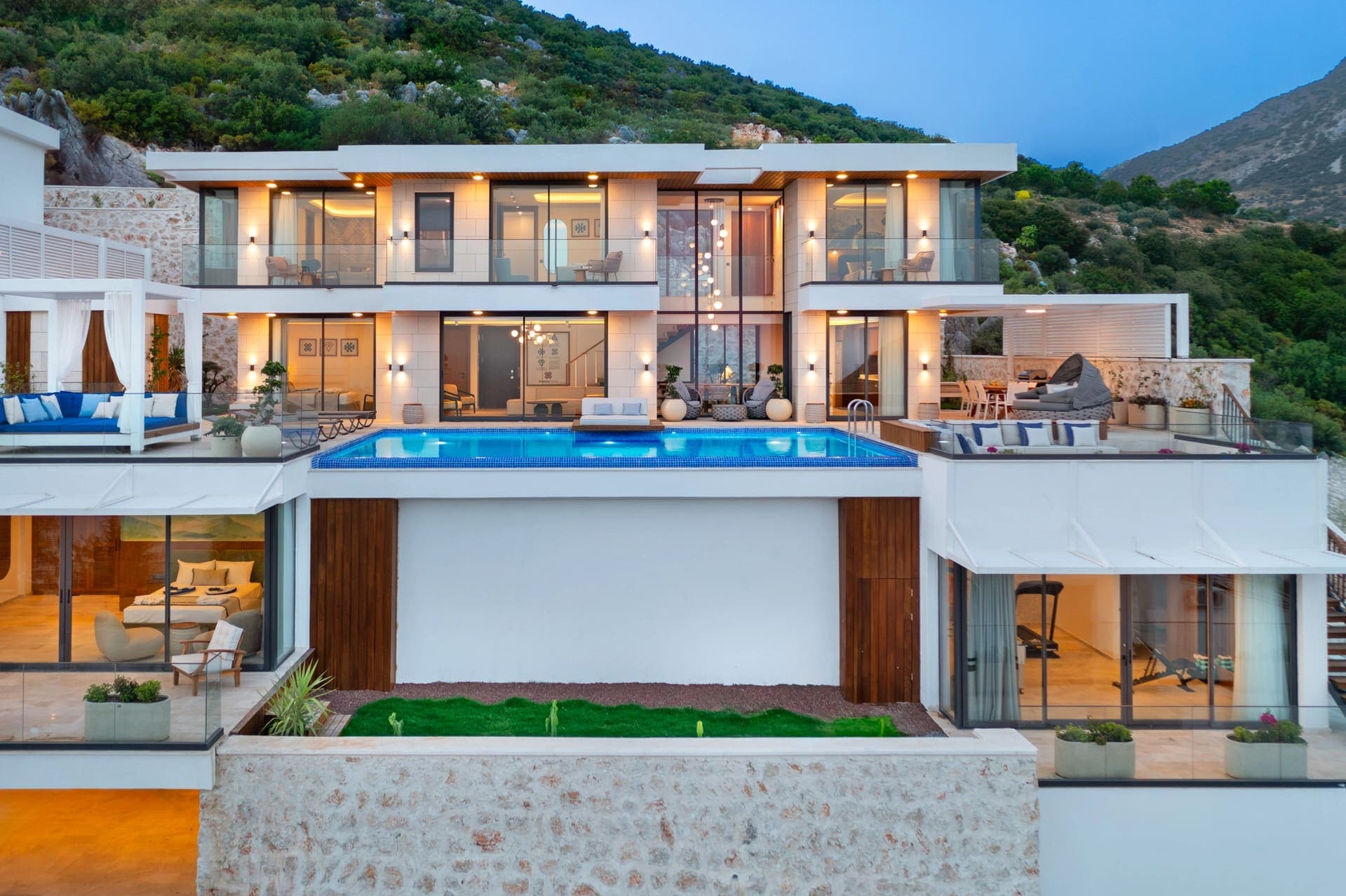 Akşam ışıkları yanan, önünde havuz ve balkonlar bulunan modern dağ villası dış cephesi.