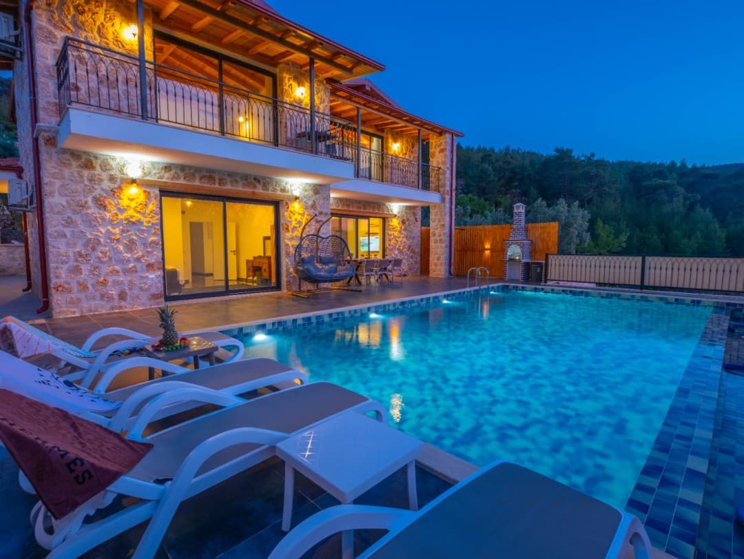Akşam ışıklandırmalı taş villa, önünde özel yüzme havuzu, şezlonglar ve bahçede barbekü alanı bulunuyor.