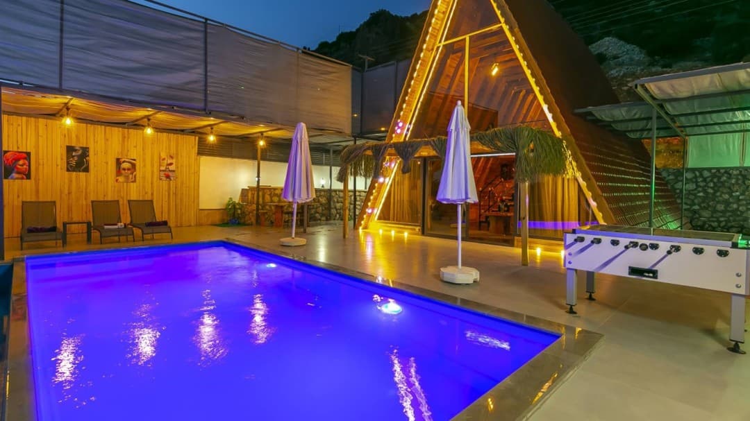 Gece aydınlatmalı havuz, yanında langırt masası ve ahşap detaylı üçgen çatılı bir villa.