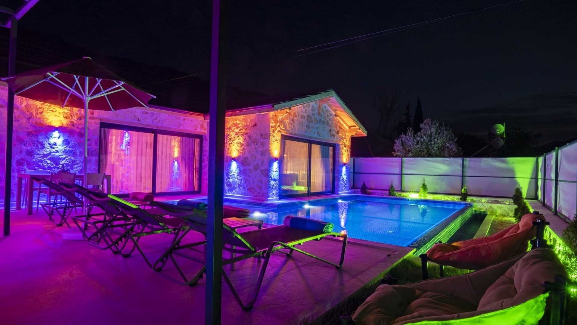 Gece vakti mor ve sarı dış cephe ışıklandırmalı taş villanın havuzu ve şezlongları.