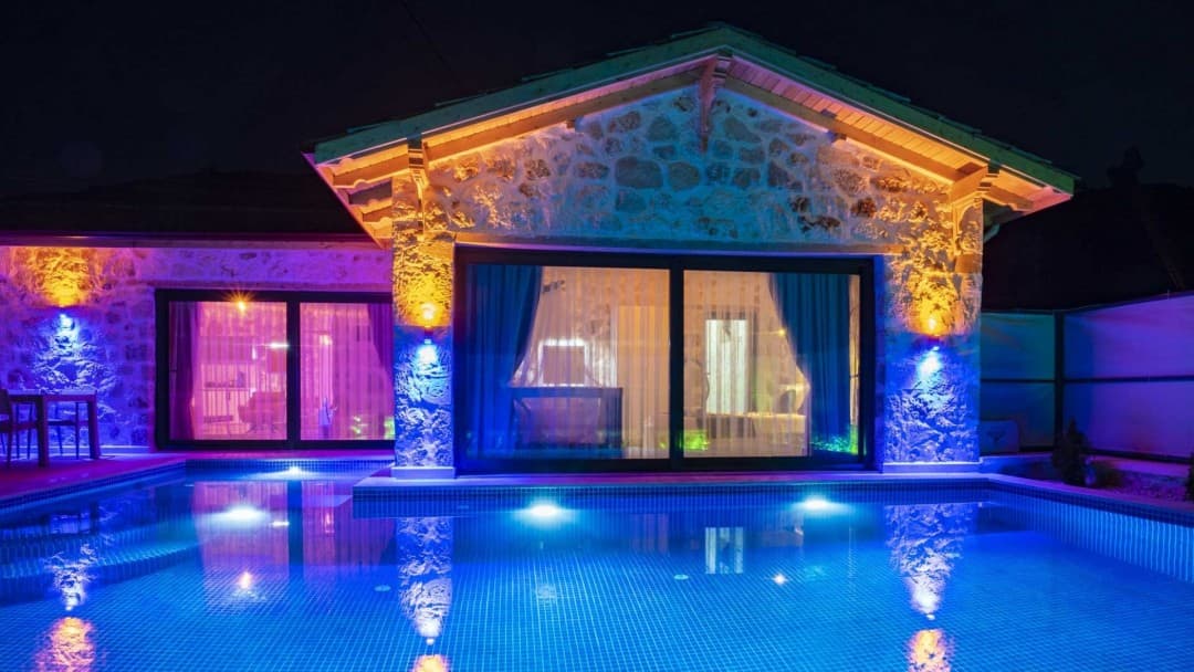 Gece renkli aydınlatmalarla ışıklandırılmış taş villa ve havuz.
