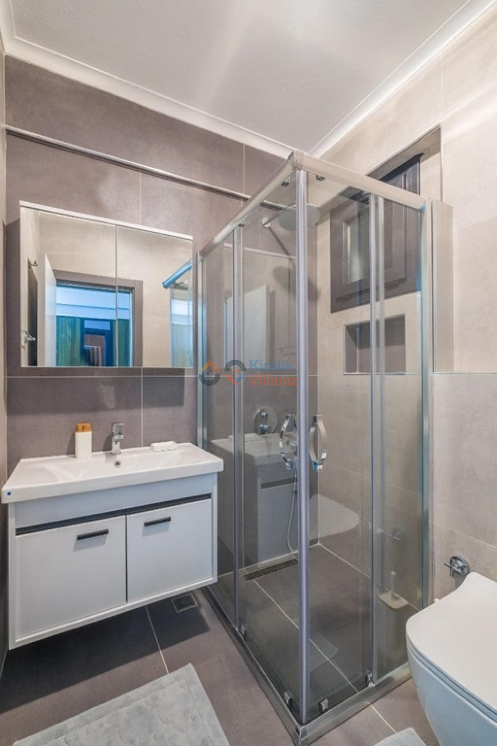 Gri tonlarında fayanslarla kaplı, cam duşakabinli ve beyaz lavabo dolaplı banyo.