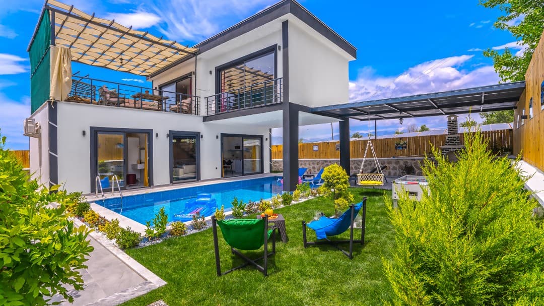 İki katlı modern villanın önünde havuz, bahçe ve renkli kamp sandalyeleri var.