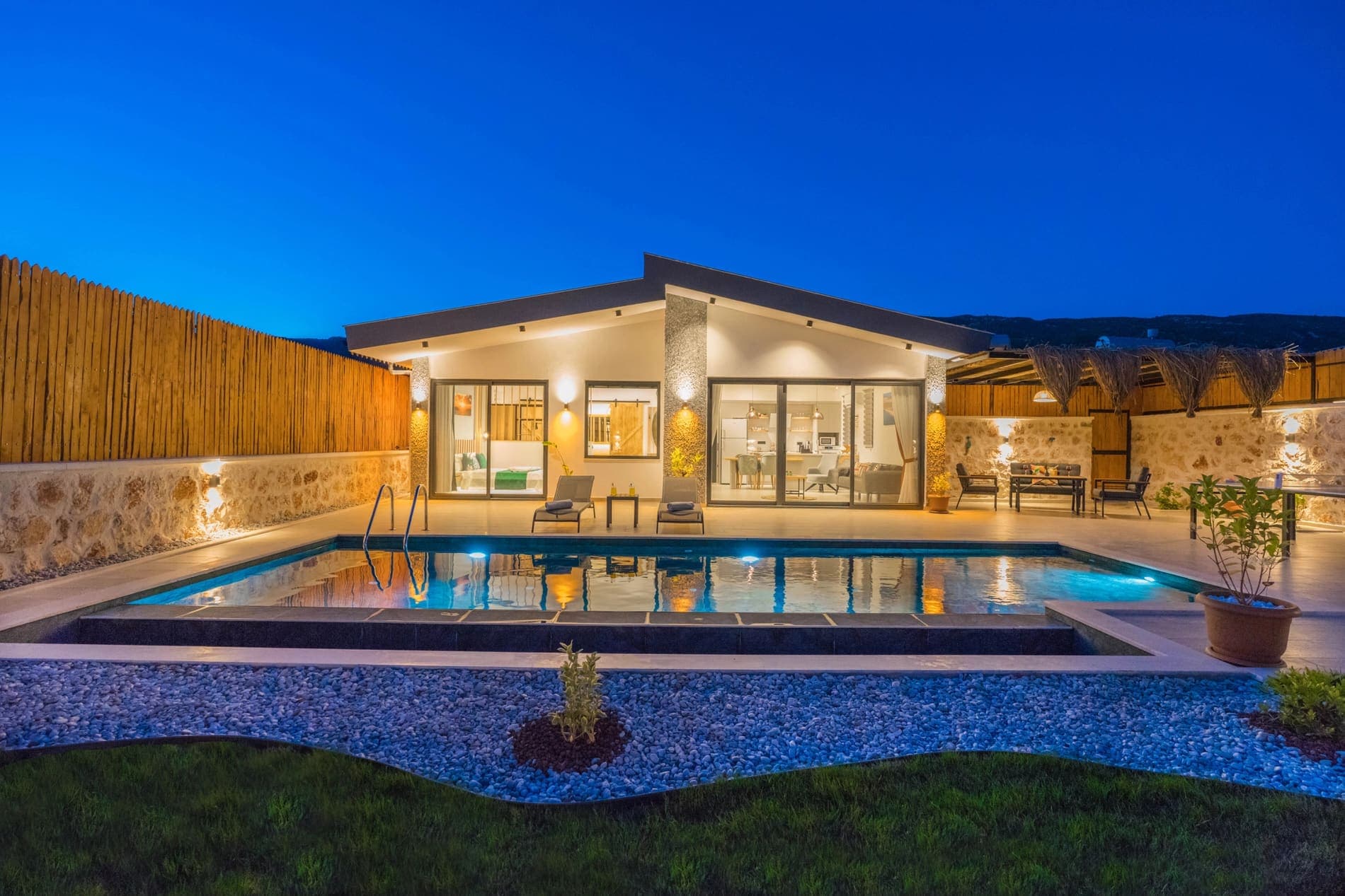 Gece aydınlatılmış modern villa, önünde dikdörtgen havuz ve bahçe mobilyaları.