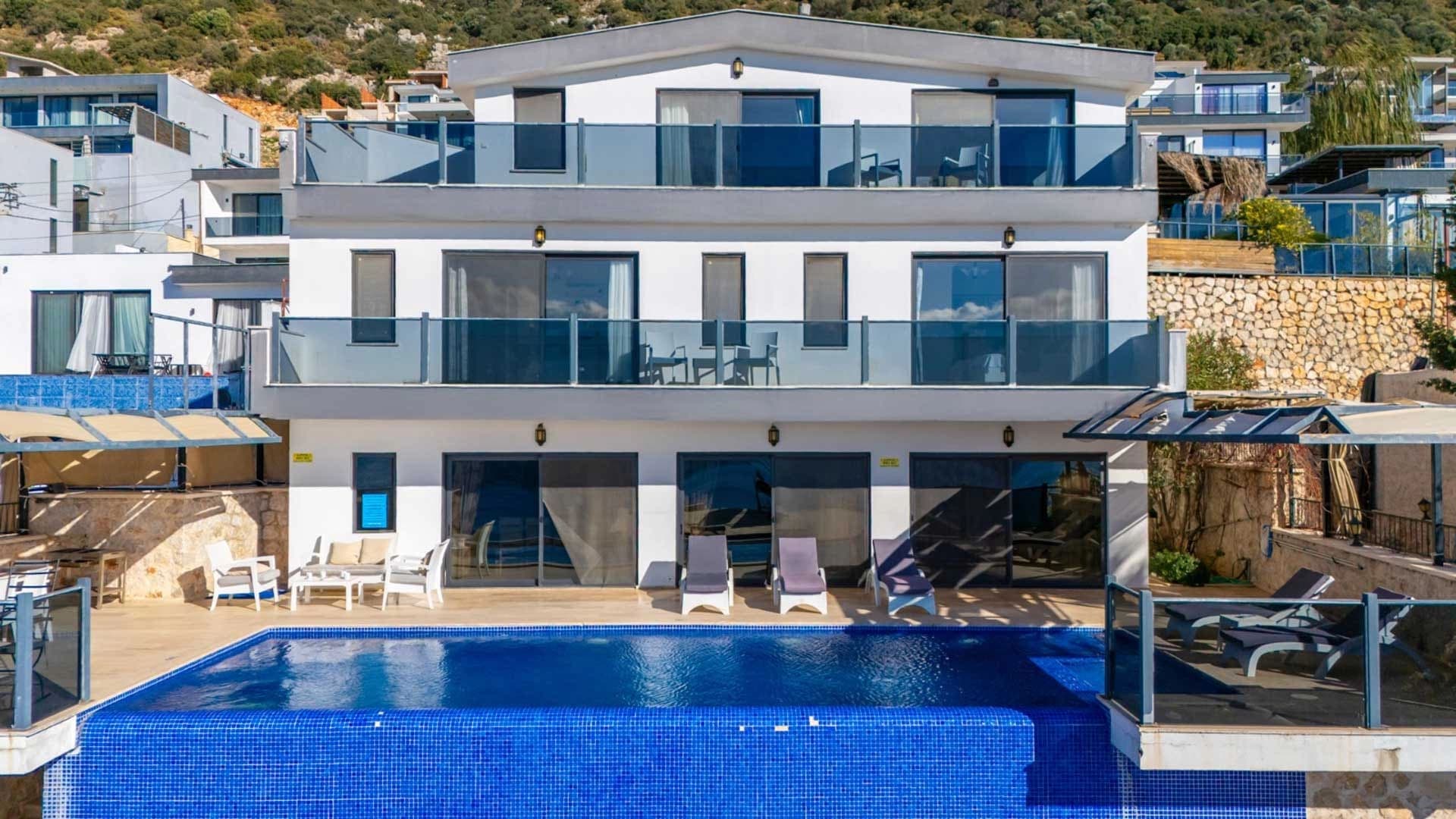 Üç katlı beyaz villa, cam balkonlar ve önünde mavi mozaikli sonsuzluk havuzu.