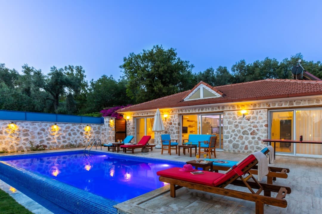 Akşam vakti aydınlatmalı havuzu ve şezlongları olan tek katlı taş villa.