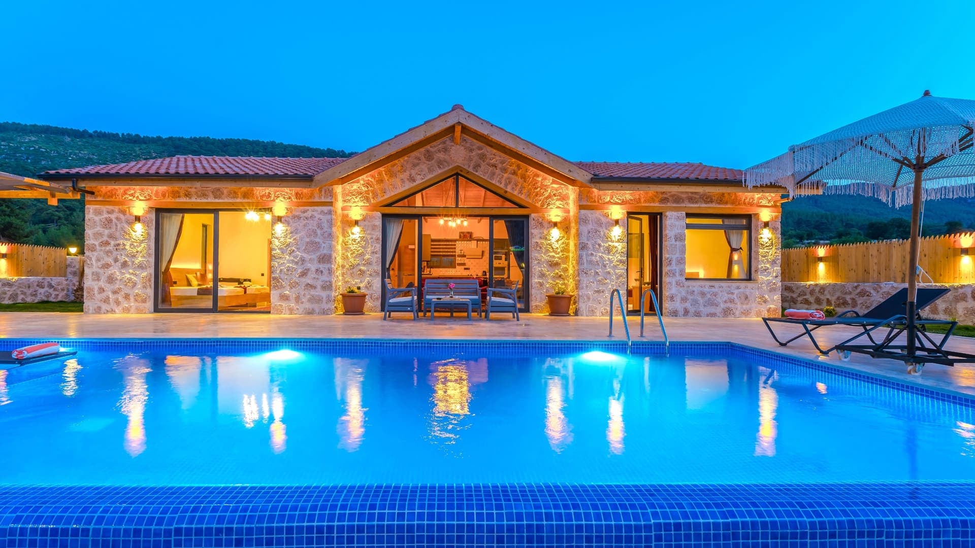 Akşam ışıklandırmasıyla aydınlatılmış taş villa, önünde mavi havuz ve şezlonglar yer alıyor.