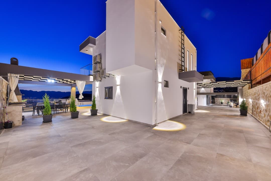 Akşam vakti aydınlatılmış modern beyaz villanın geniş terası, saksı bitkileri ve oturma grubu.