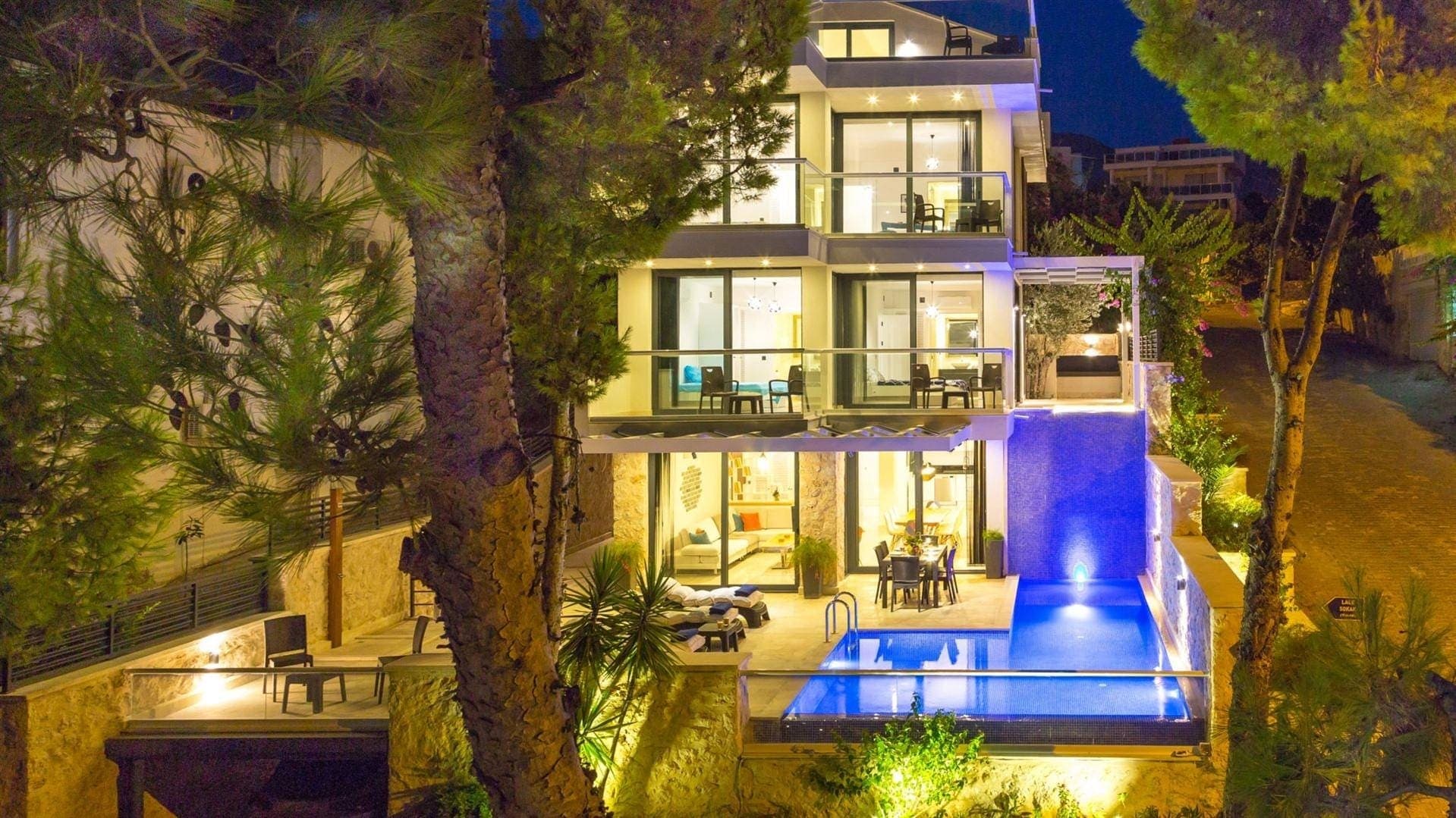 Gece aydınlatılmış, katlı, modern mimariye sahip villa ve önündeki ışıklandırılmış yüzme havuzu.