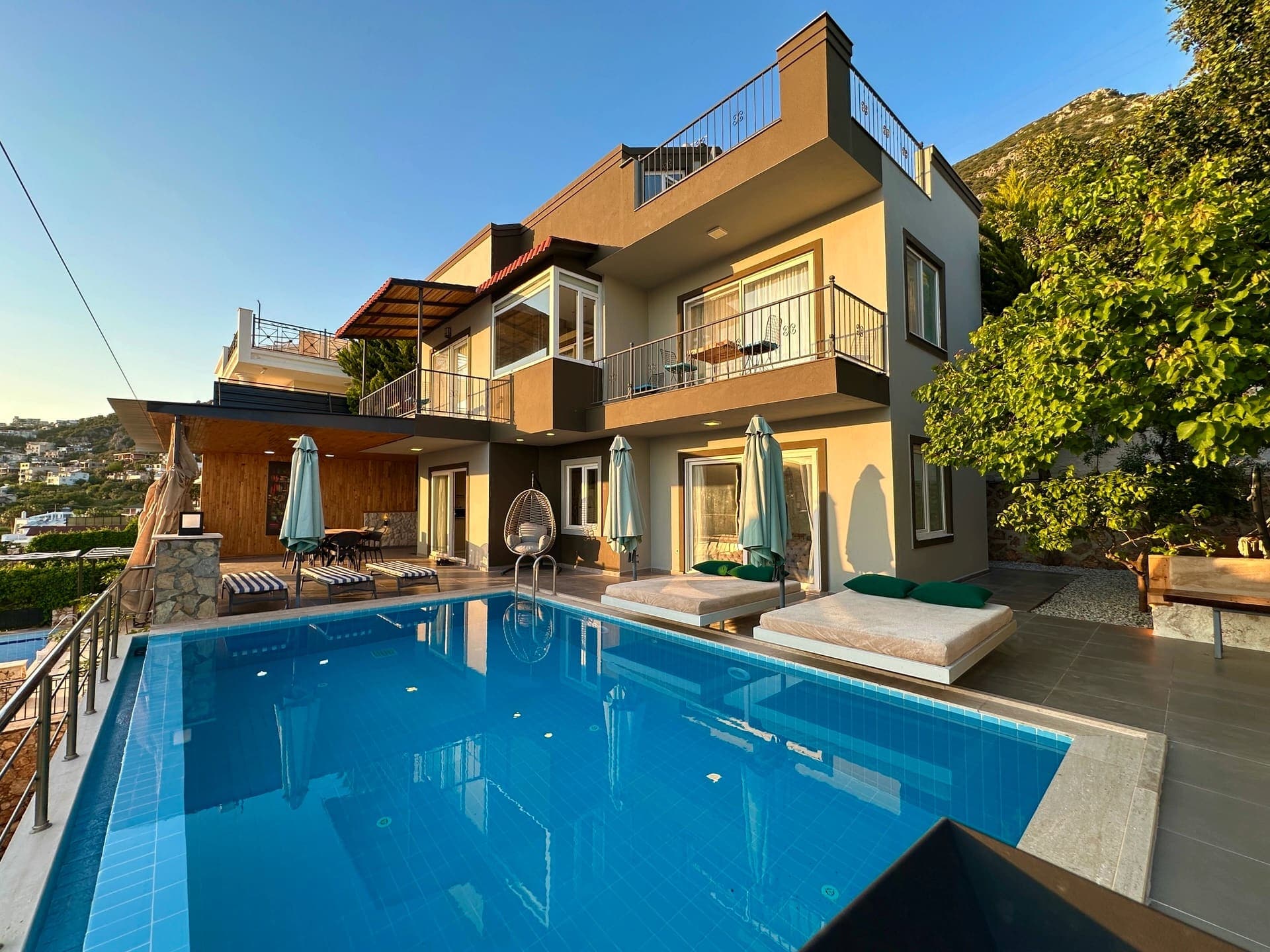 Gün batımı ışığında modern villa cephesi, önünde havuz, şezlonglar ve asma bahçe koltuğu.