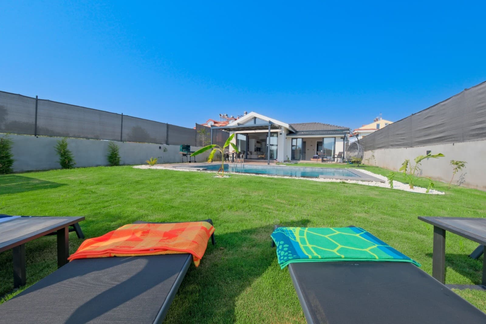 Ön planda şezlonglar, arkada çimli bahçe ve havuzlu villa bulunuyor.