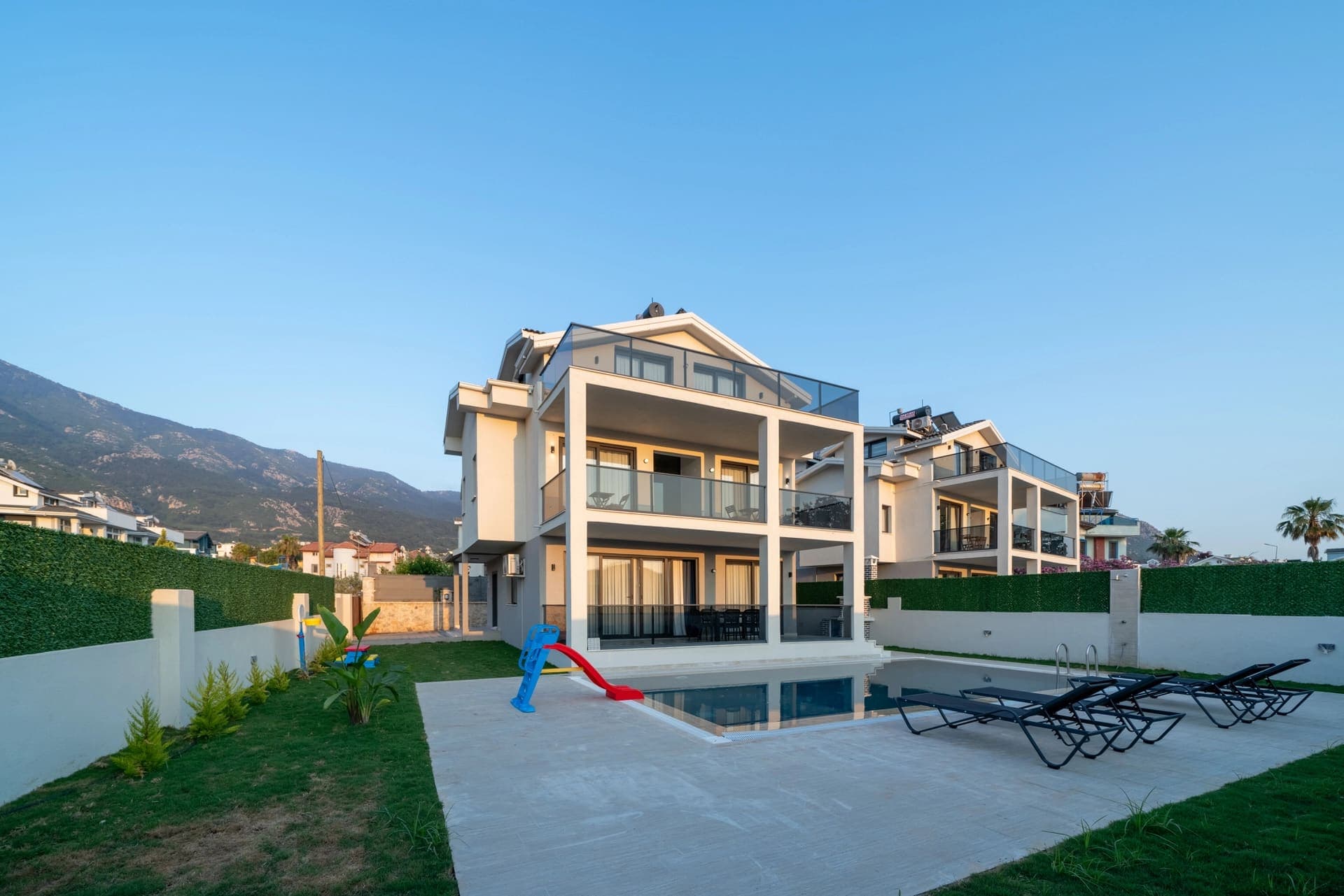 Üç katlı modern villa, önünde yüzme havuzu, şezlonglar ve çocuk kaydırağı bulunan bahçesi.