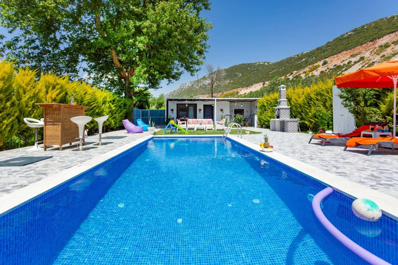Bahçesinde yüzme havuzu, bar masası, şezlonglar ve taş barbekü bulunan villa.