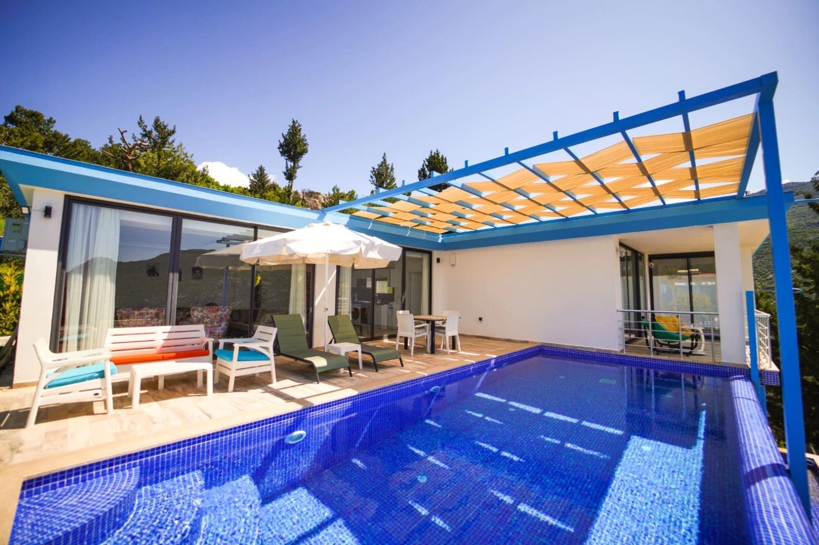 Mavi gölgelikli terası, yüzme havuzu ve dış mekan oturma grubu olan beyaz villa.