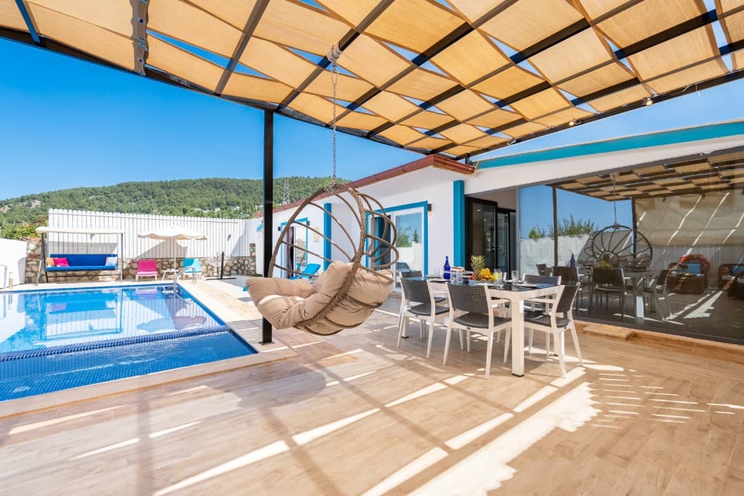 Güneşlikli terasta asılı salıncak, yemek masası ve yanında özel yüzme havuzu bulunan villa dış alanı.