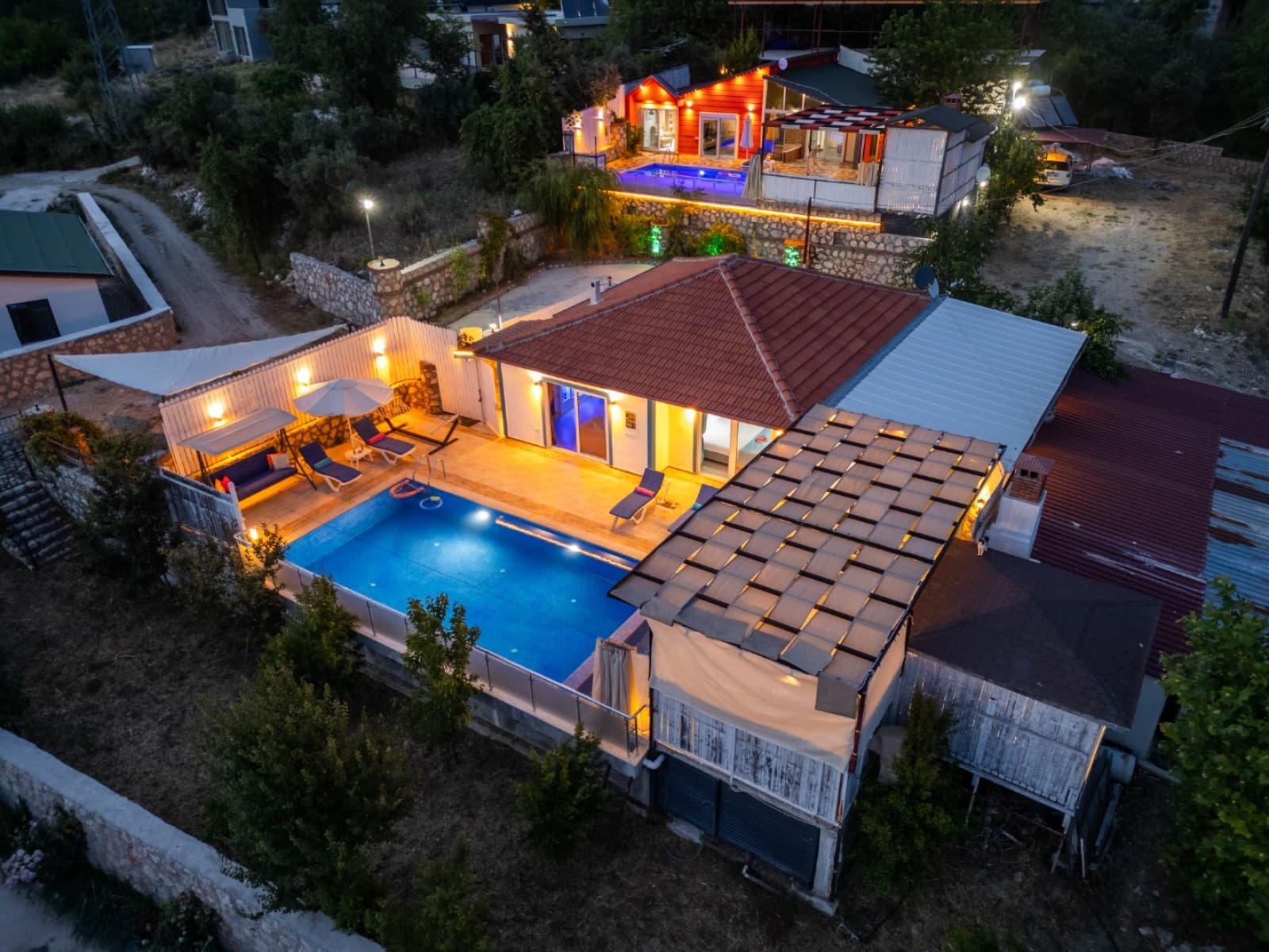 Havuzlu ve kiremit çatılı villanın akşam havadan çekilmiş ışıklı görüntüsü.