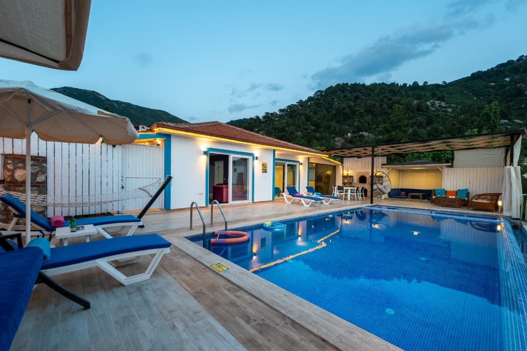 Akşam vakti havuz başı şezlongları, şemsiyesi ve dağ manzaralı tek katlı villa.