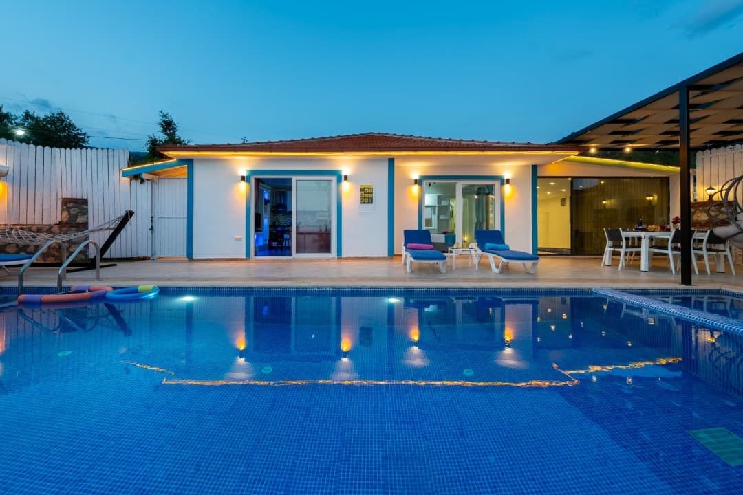 Akşam vakti aydınlatılmış havuzun arkasındaki beyaz tek katlı villa ve şezlonglar.