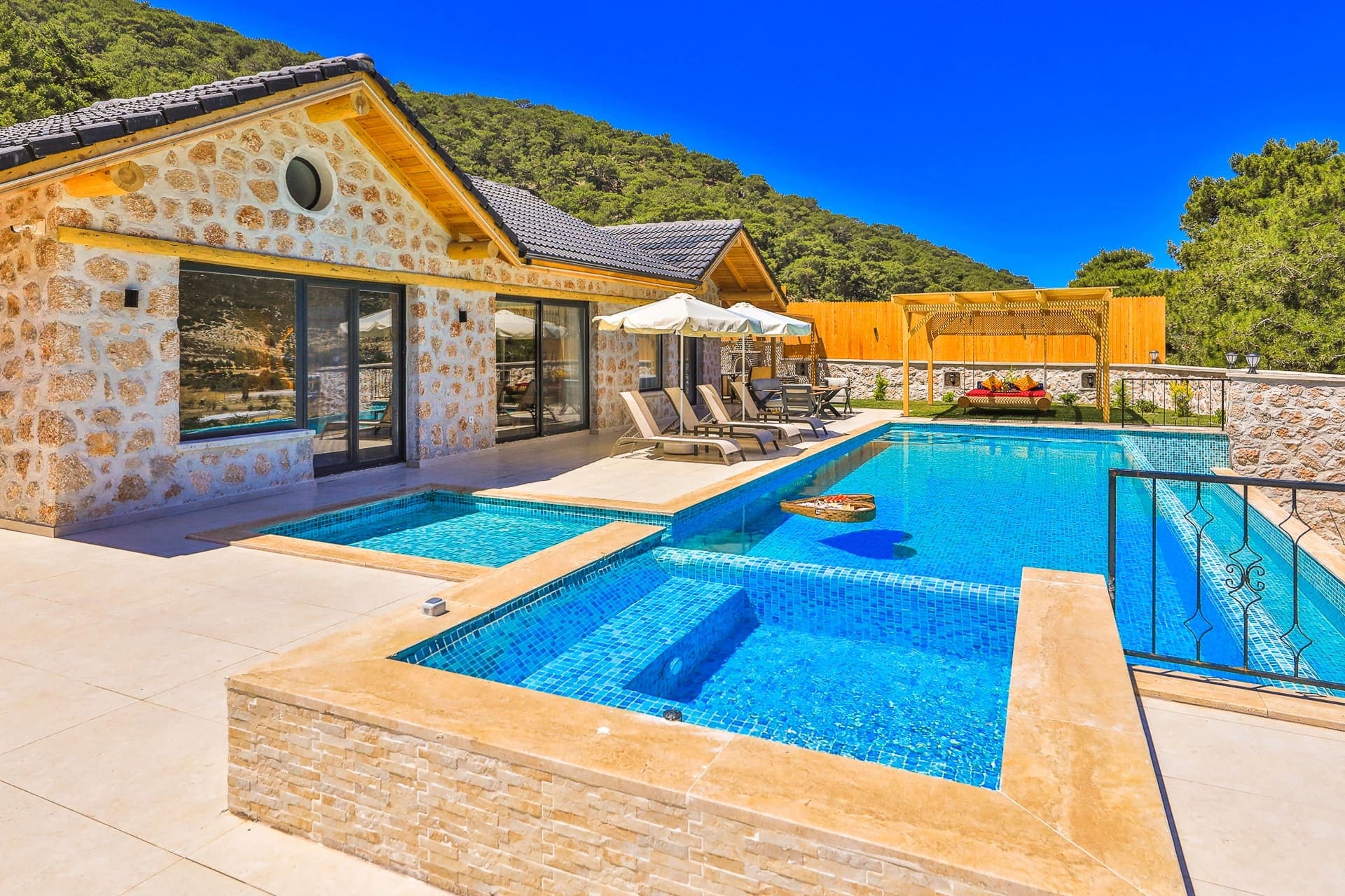Taş kaplama villa, önünde şezlonglar, güneş şemsiyeleri ve kademeli yüzme havuzu.