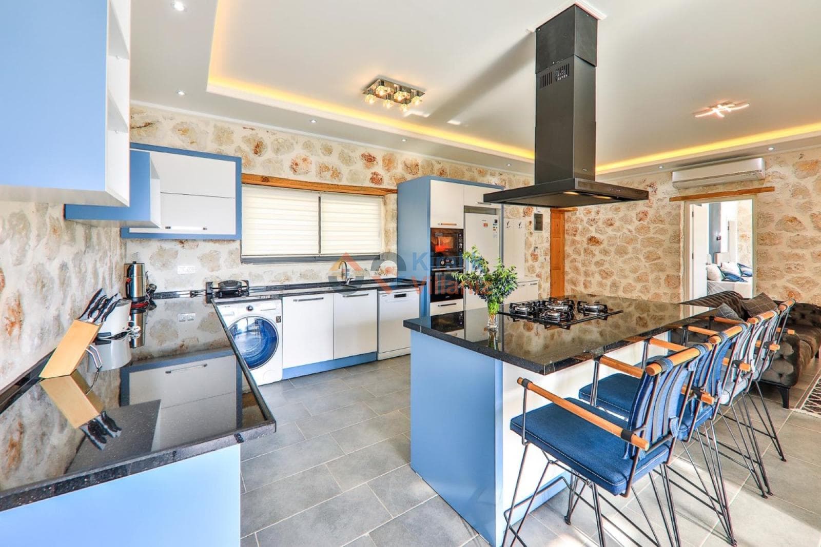 Taş duvarlı, mavi dolaplı, ada mutfaklı ve çamaşır makineli modern mutfak ve oturma alanı.