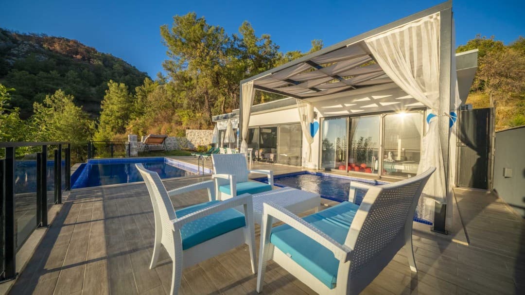 Ön planda beyaz oturma grubu, yanında havuz ve güneşlikli terası olan orman manzaralı tek katlı villa.