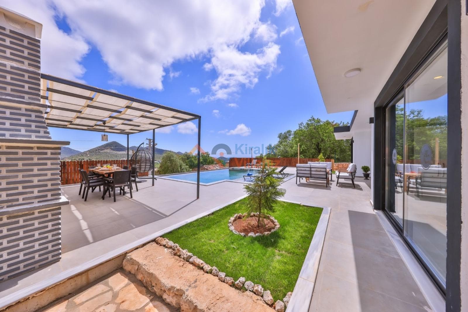 Havuz kenarında yemek masası, oturma grubu ve küçük bir bahçe alanı bulunan villa terası.