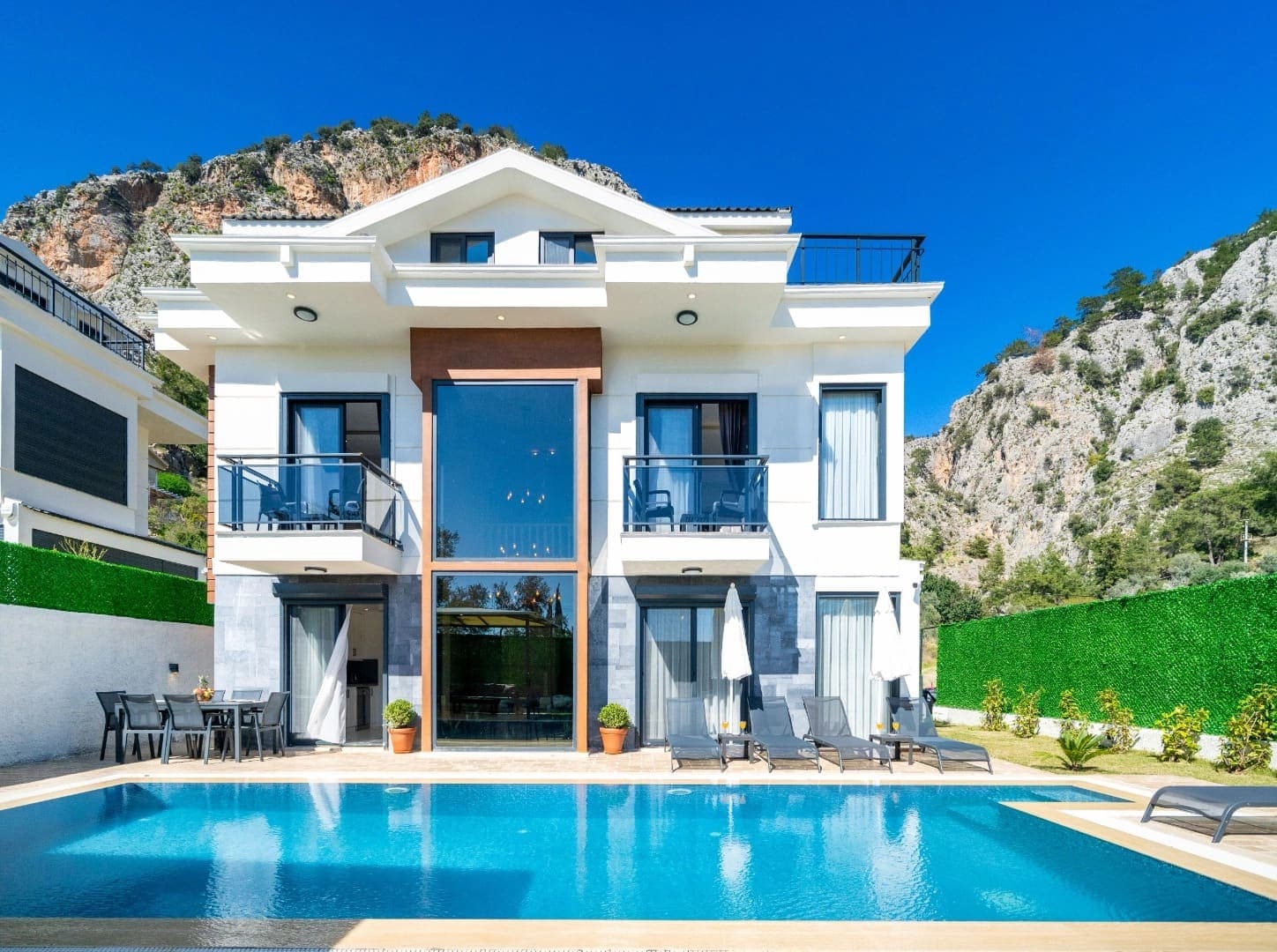 Dağ manzaralı, önünde havuz ve şezlonglar bulunan beyaz dış cepheli villa.