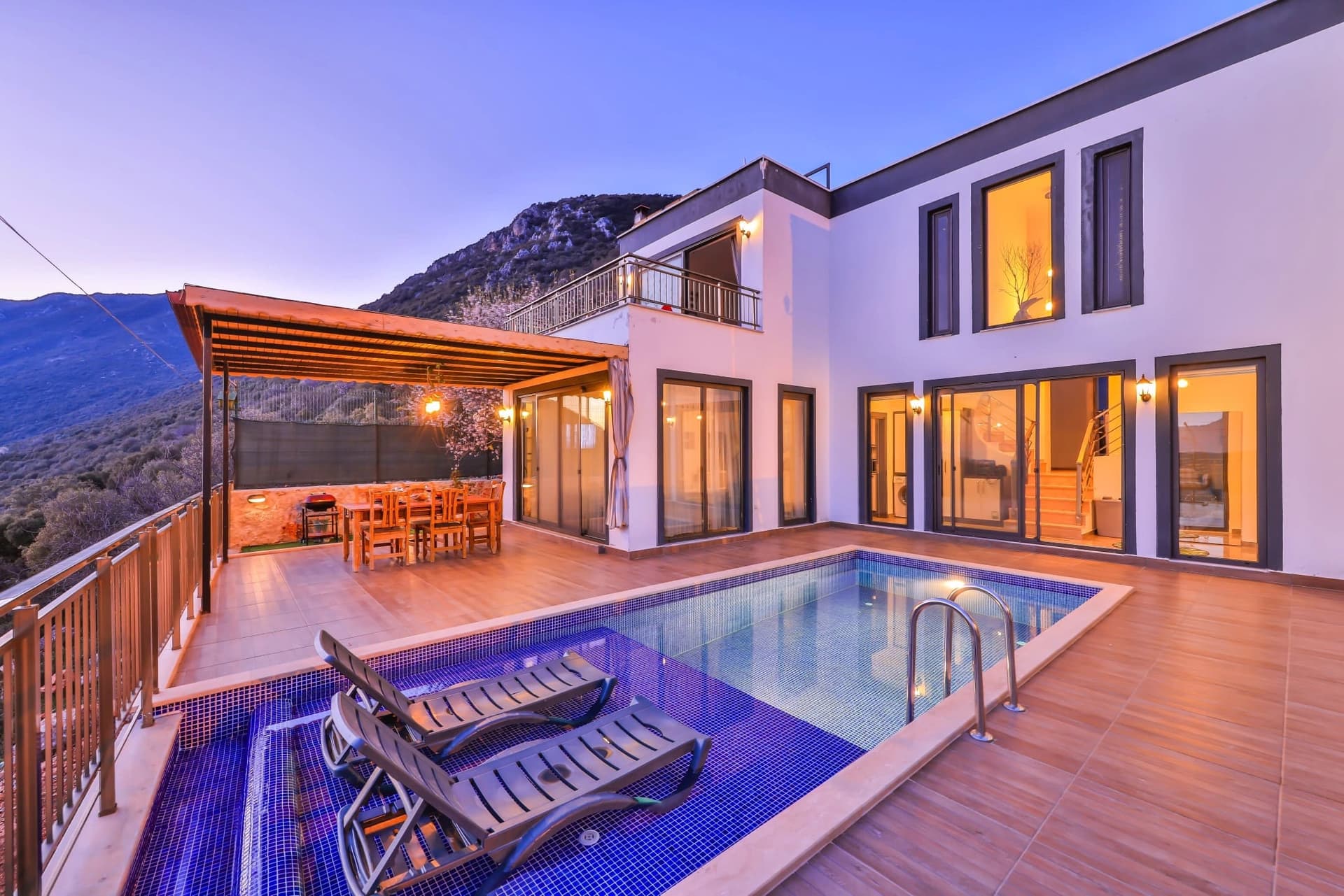 Akşam vakti aydınlatılmış havuz, şezlonglar ve gölgelikli veranda alanı bulunan beyaz dış cepheli villa.
