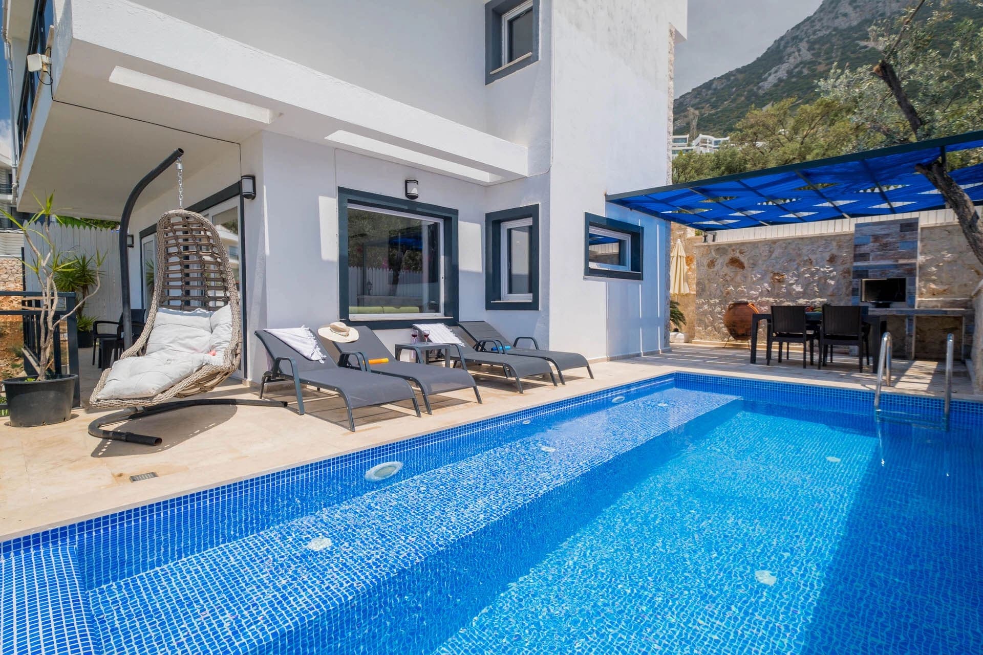 Havuz kenarında şezlonglar, salıncak ve gölgelikli yemek alanı bulunan villa dış cephesi.