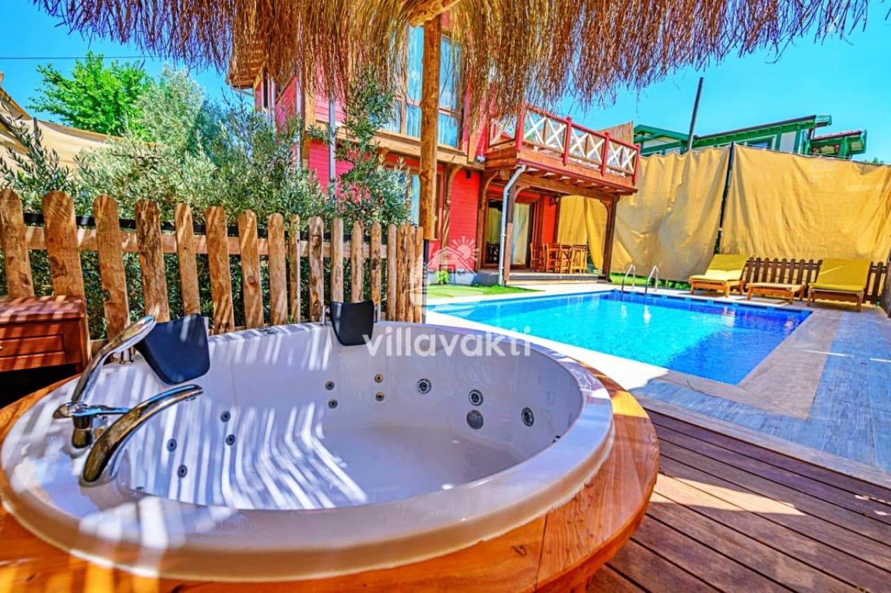 Ahşap güverte üzerinde yuvarlak jakuzi, arka planda yüzme havuzu ve kırmızı ahşap villa.