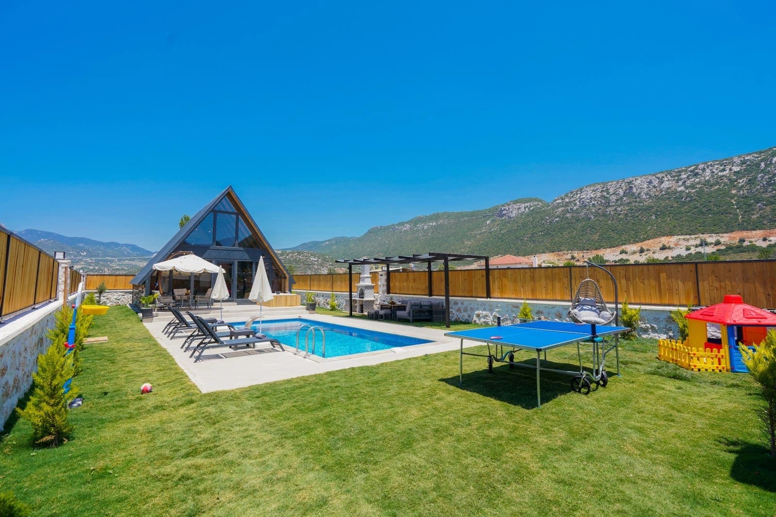 Bahçesinde havuz, masa tenisi, çocuk oyun alanı ve üçgen çatılı bungalov ev bulunan geniş alan.