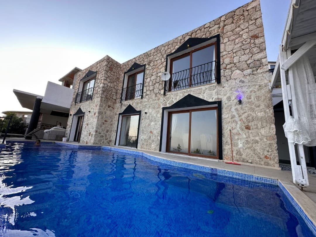 Taş kaplama dış cepheli, balkonlu ve önünde mavi fayanslı yüzme havuzu bulunan iki katlı villa.