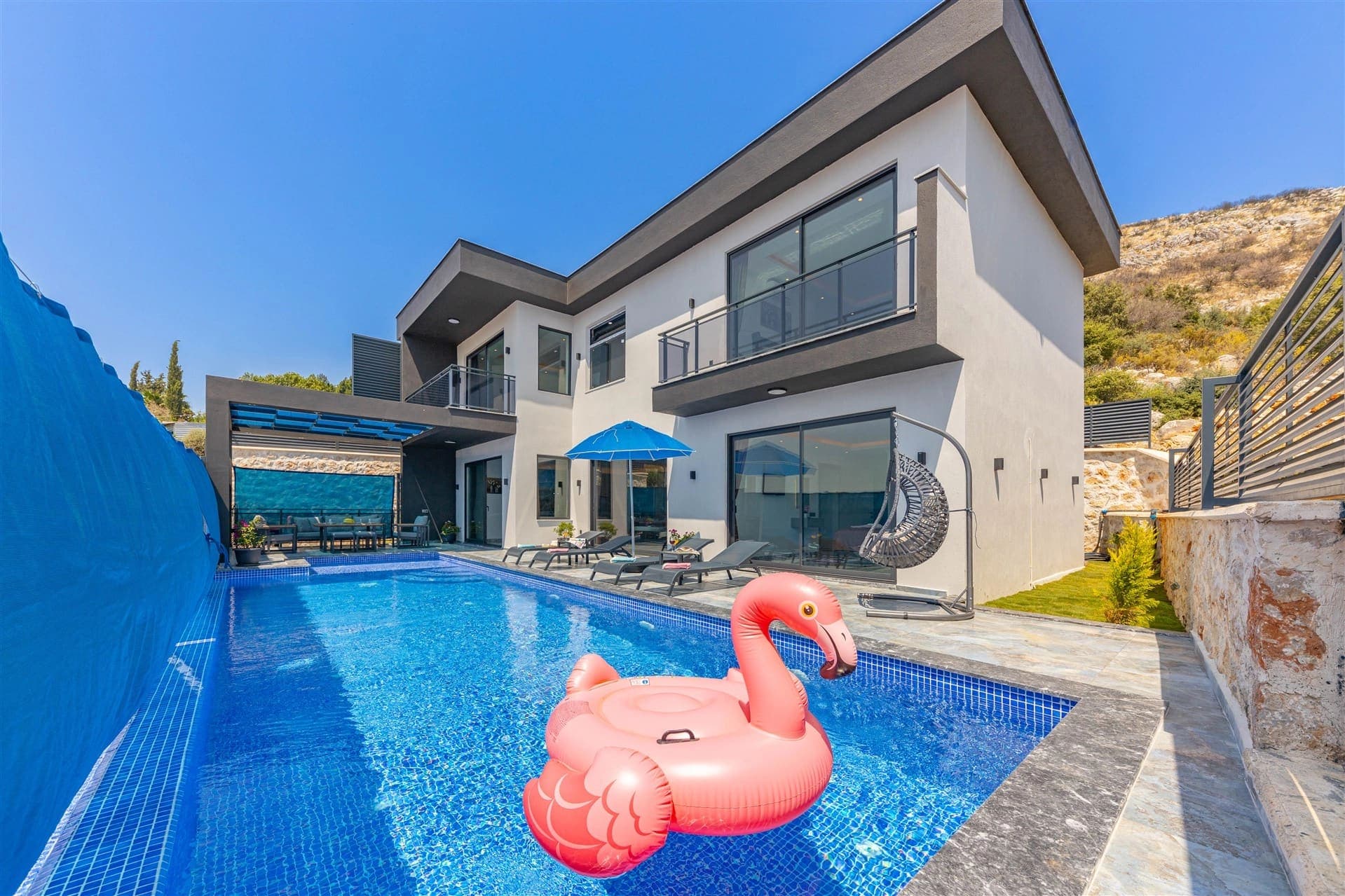 Modern villanın önünde pembe flamingo deniz yatağı bulunan yüzme havuzu ve güneşlenme alanı var.