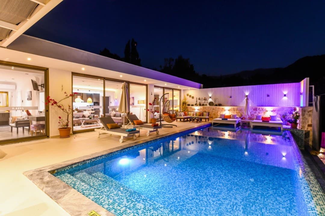 Gece vakti havuz başı aydınlatmalı modern villanın dış cephesi ve şezlonglar.