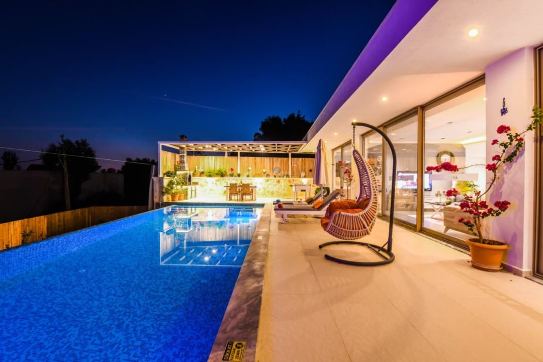 Gece aydınlatmalı havuz kenarında salıncak, şezlonglar ve arka planda yemek alanı bulunan villa terası.