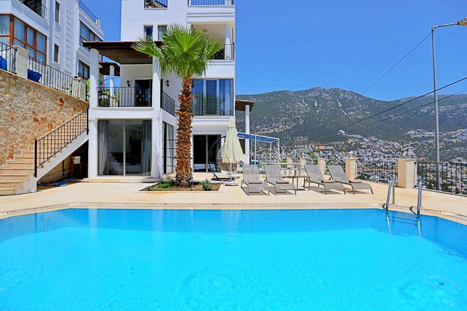 Mavi yüzme havuzu, şezlonglar, palmiye ağacı ve arka planda dağ manzaralı beyaz bir villa.