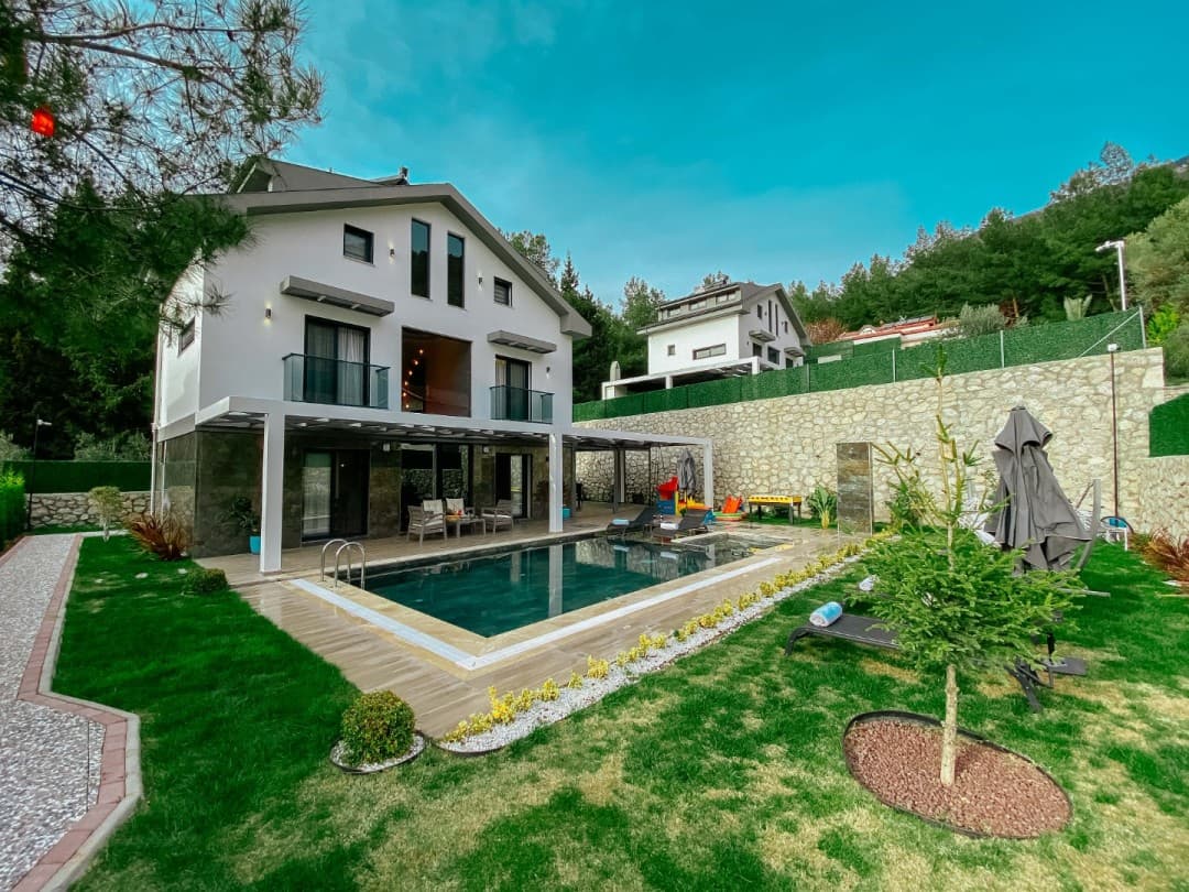 Gündüz saatlerinde havuzlu, çim bahçeli ve taş duvarlı iki katlı beyaz villa.
