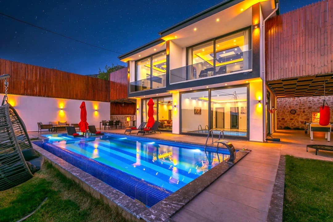 Akşam vakti aydınlatılmış iki katlı modern villanın önünde havuz ve şezlonglar var.