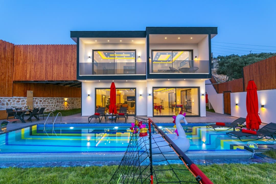 Akşam ışıklandırmalı, havuzlu ve bahçesinde deniz atı şeklinde şişme bot bulunan modern iki katlı villa.