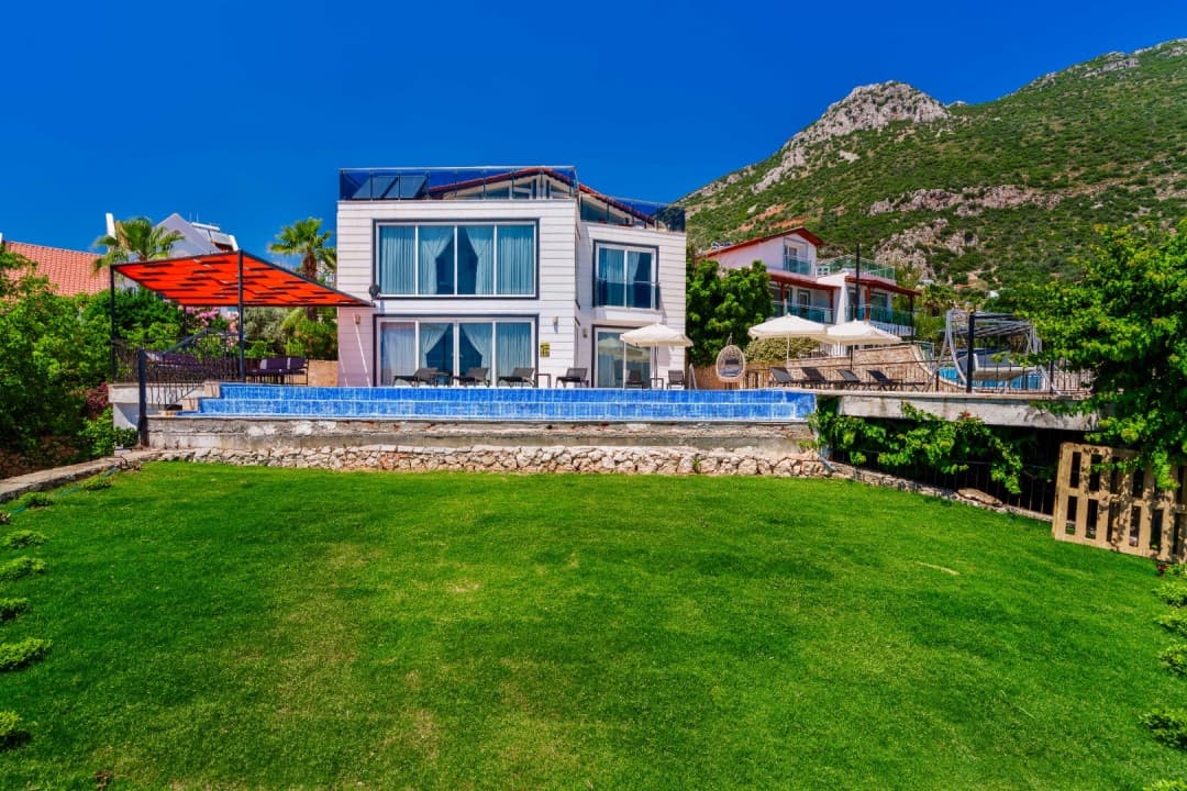 Ön planda çim bahçe, arkasında sonsuzluk havuzu, şezlonglar ve dağ manzaralı beyaz katlı bir villa.