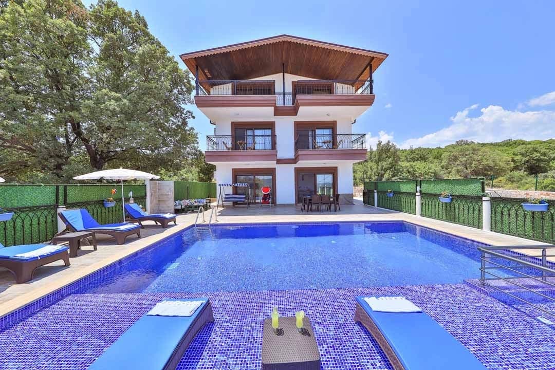 Üç katlı, balkonlu villa binası ve önünde mavi fayanslı yüzme havuzu ile şezlonglar.