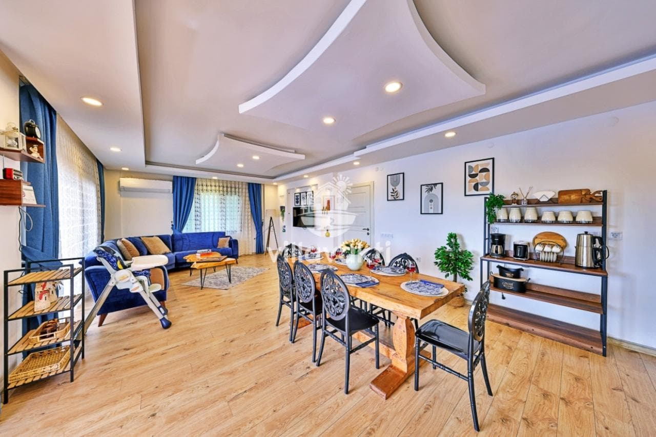 Modern salon ve yemek odasında ahşap yemek masası, mavi koltuk takımı ve açık mutfak rafı bulunuyor.