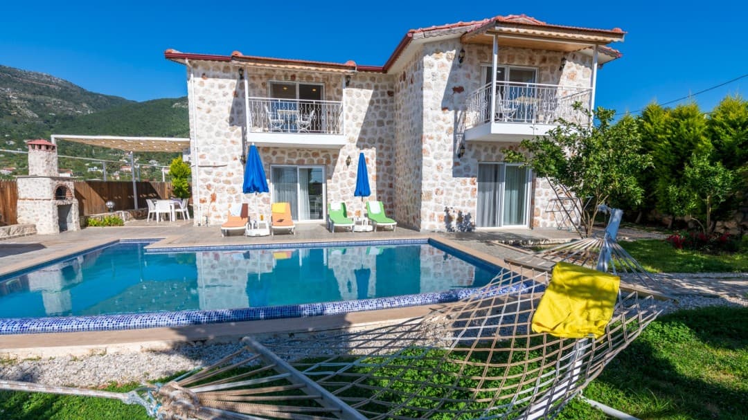 Önünde yüzme havuzu ve şezlonglar olan iki katlı taş villa ve çim alanda hamak.
