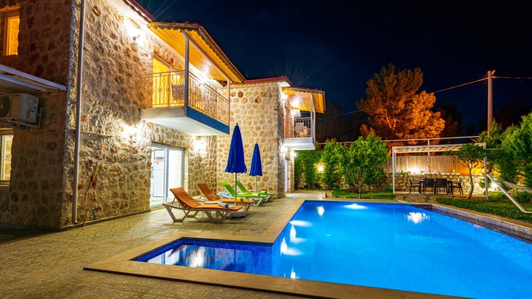 Gece vakti ışıklandırılmış taş villanın önünde havuz ve şezlonglar yer alıyor.