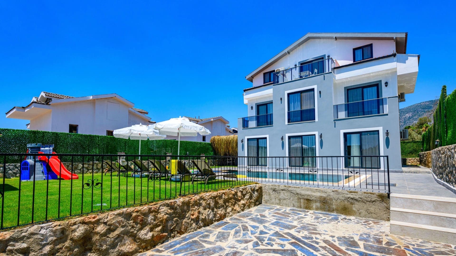 Çocuk oyun parkı ve şezlongların bulunduğu bahçeye sahip gri renkli villa dış cephesi.