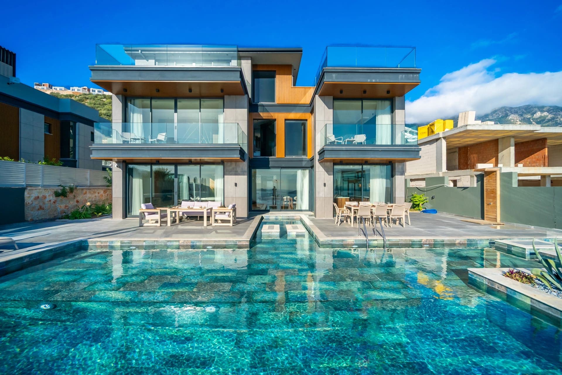 Modern mimariye sahip villa, önünde basamaklı havuz ve güneşlenme terasında yemek masası bulunuyor.