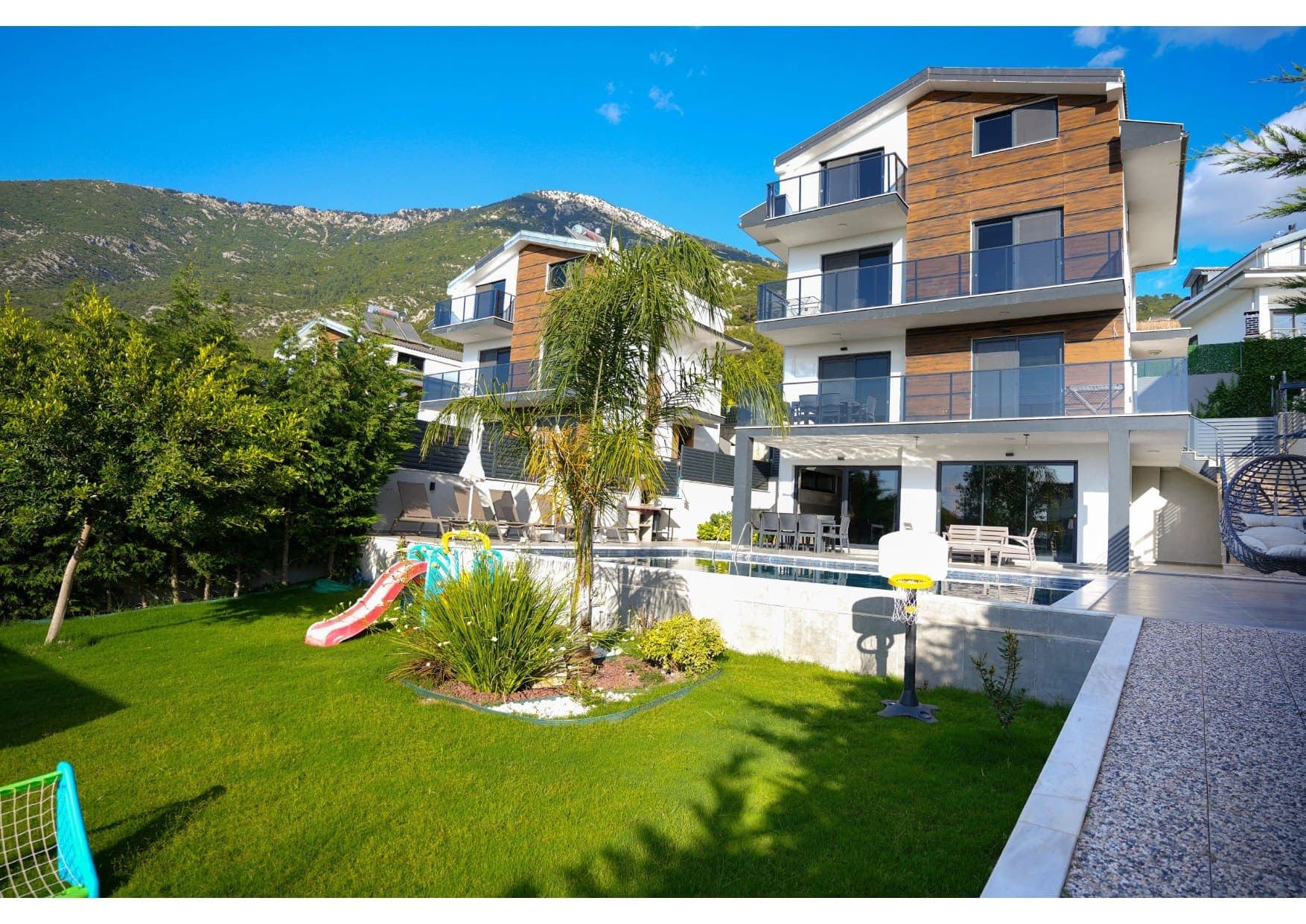 Üç katlı modern villa, bahçesinde çocuk oyun alanı, havuz ve dağ manzarası ile dikkat çekiyor.