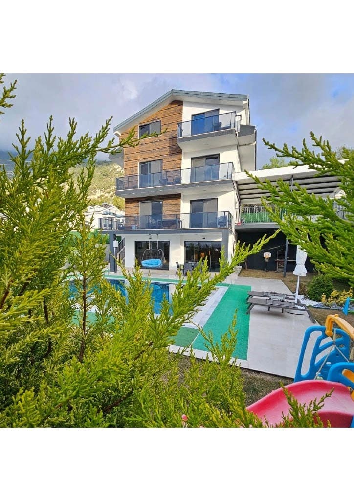 Ağaçların arasından görünen havuzlu, üç katlı modern villa ve bahçesindeki çocuk oyun alanı.