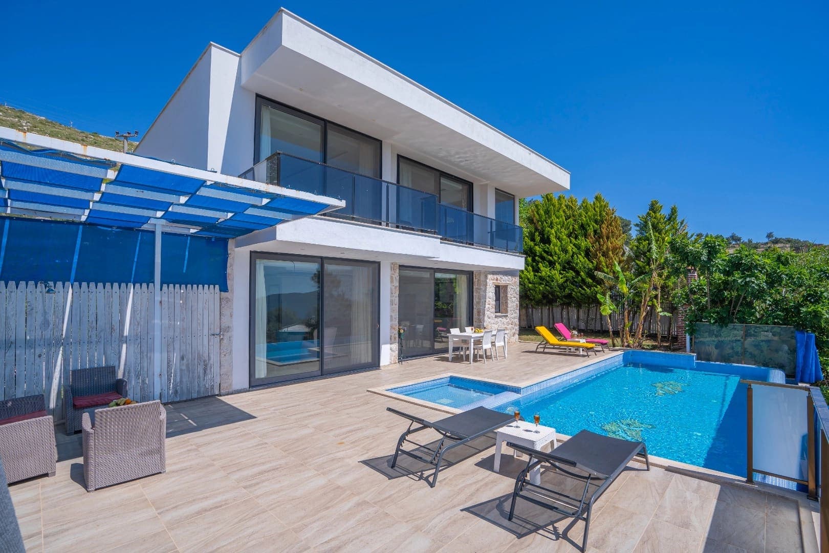 İki katlı beyaz modern villa, önünde özel havuz, şezlonglar ve yemek masası bulunan geniş bir teras.